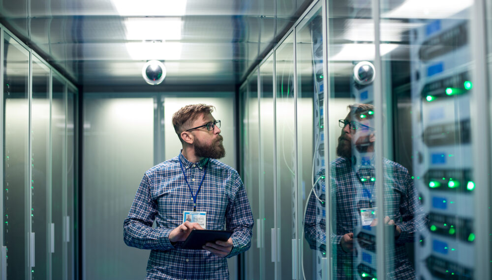 Wide shot of casual man with tablet walking among server racks in data center room looking at hardware